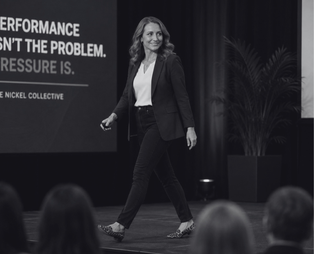 A woman with wavy hair wearing a blazer, white shirt, black pants, and animal print shoes is walking on stage during a presentation or lecture, with a large screen and audience in the background.