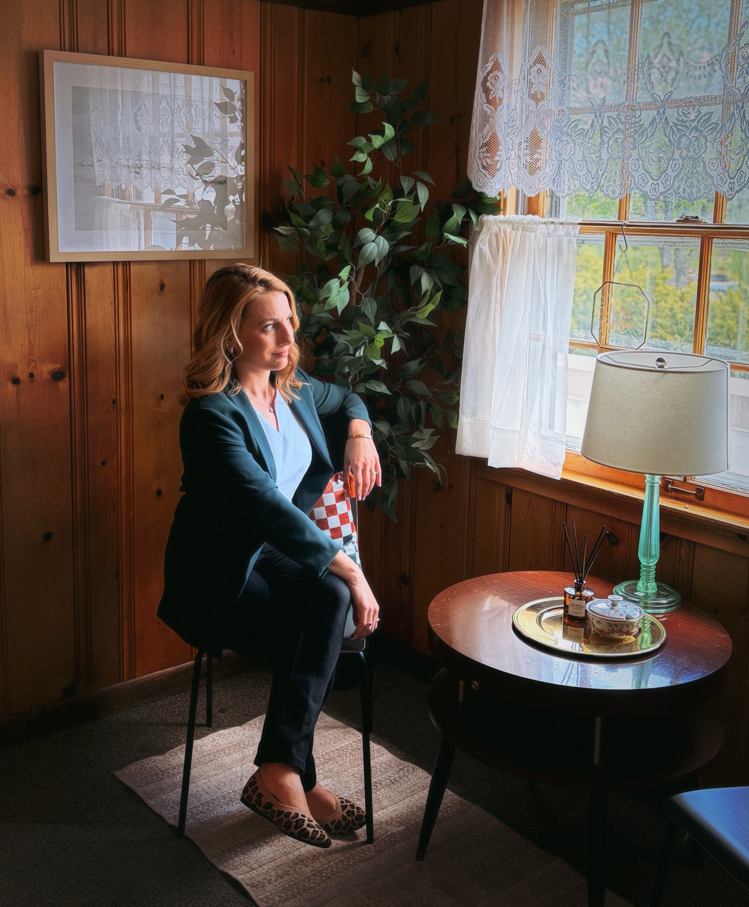 A woman with blonde hair sitting on a black chair in a wood-paneled room, near a round black table with decorative items, a lamp, and windows with lace curtains, sunlight streaming in.