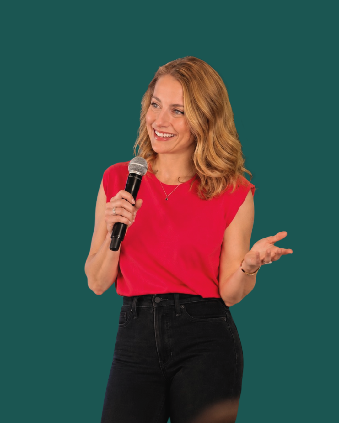 A woman with shoulder-length blonde hair wearing a red sleeveless top and black jeans, holding a microphone and speaking with hand gestures against a teal background.