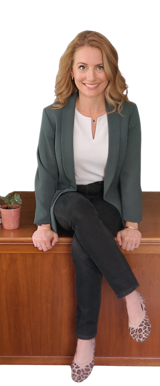 A smiling woman with blonde hair, wearing a gray blazer, white top, black pants, and leopard print heels, sitting on a wooden cabinet next to a small potted plant.