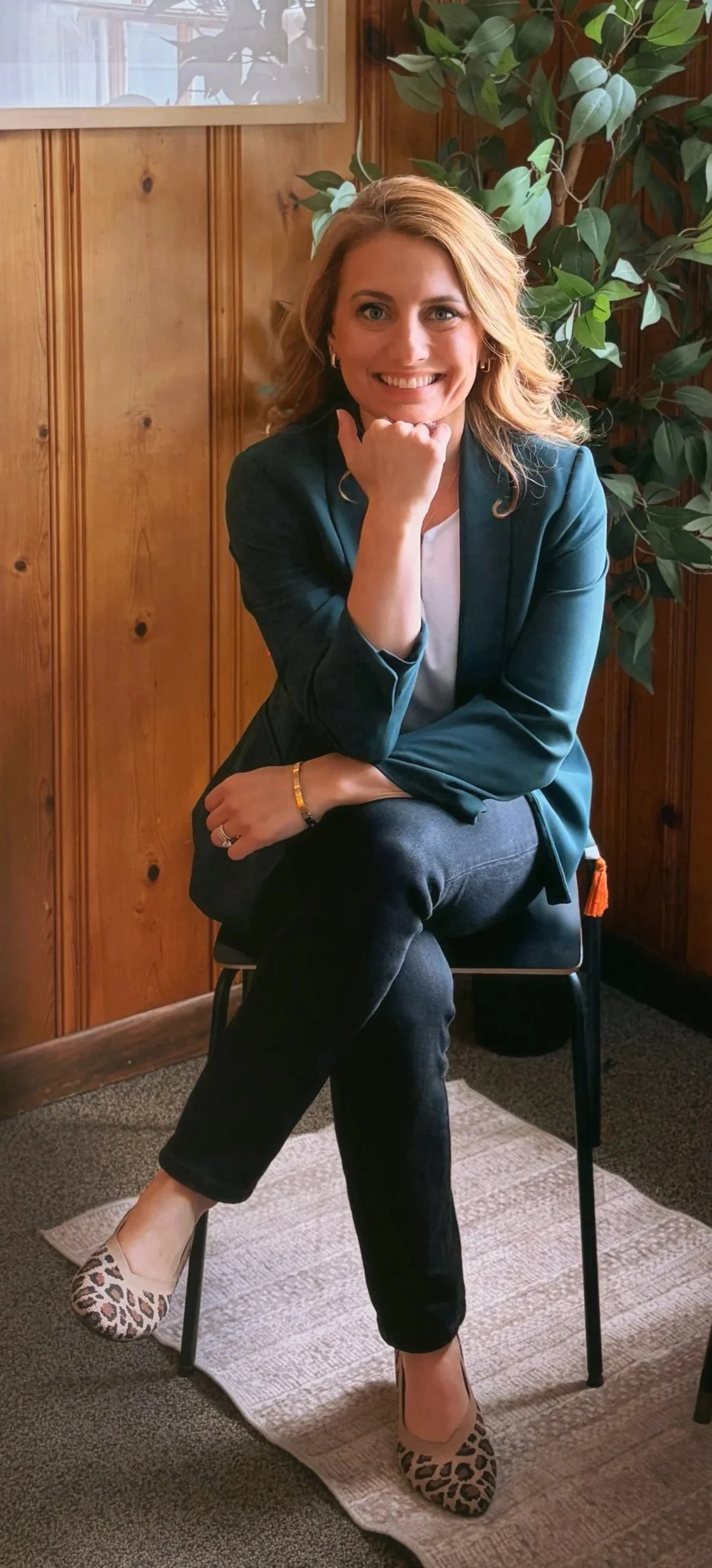 A woman with blonde hair, wearing a teal blazer, white shirt, black jeans, leopard print flats, and jewelry, sitting on a black chair with her right foot crossed over her left knee, in a room with wooden walls, a plant behind her, and a window above with light coming through.