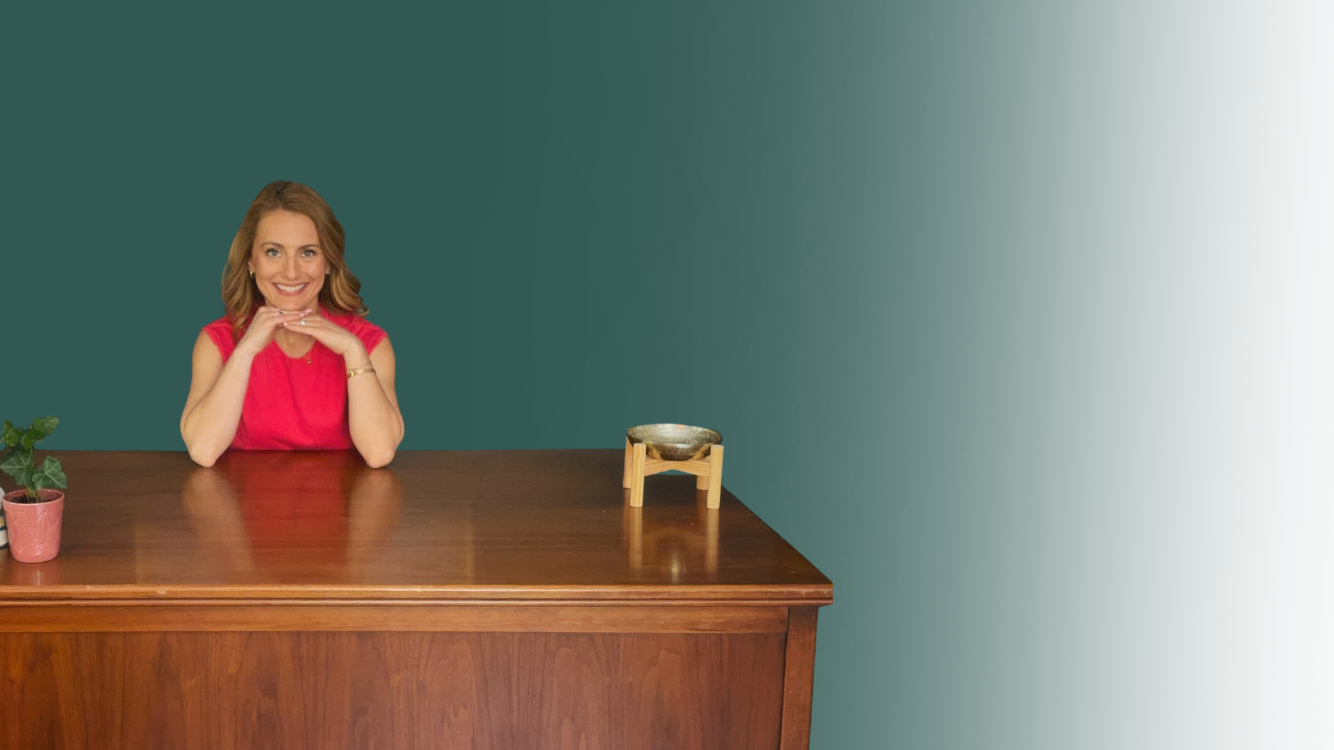 A woman in a red sleeveless top smiling with her hands under her chin, seated at a wooden desk with a potted green plant and a decorative bowl on a small wooden stand, against a teal and white gradient wall background.