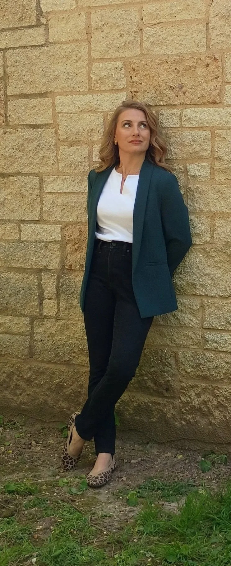 A woman with wavy blonde hair, wearing a dark green blazer over a white top, black jeans, and leopard print shoes, standing outdoors against a beige brick wall with grass at her feet.