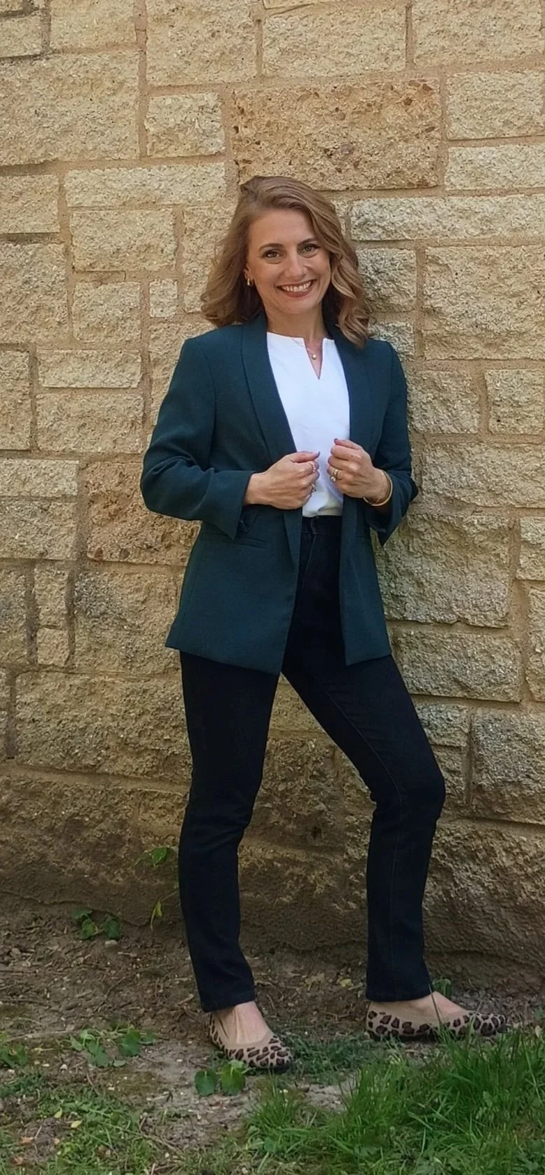 A woman with blonde hair in loose waves, wearing a teal blazer over a white blouse, black pants, and leopard print shoes, standing outdoors against a textured beige brick wall. She is smiling and holding her blazer lapels.