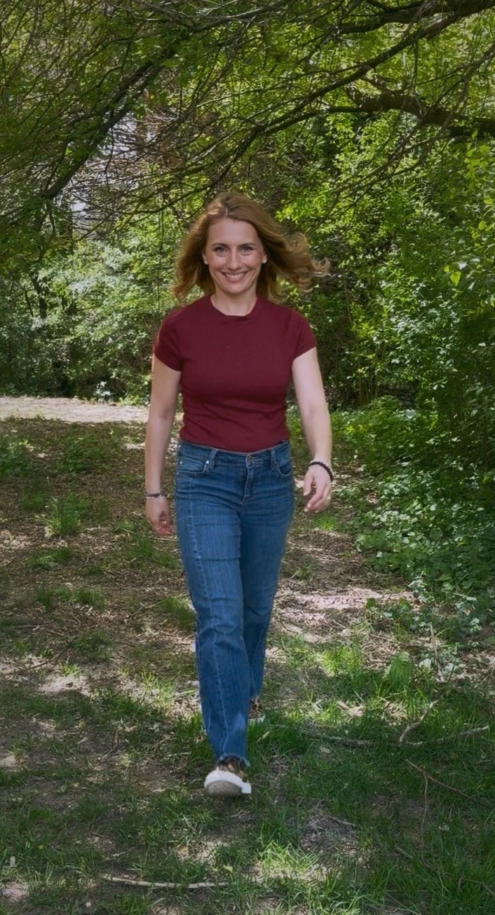 A woman with shoulder-length wavy hair walking on a dirt trail through a lush green forest, wearing a maroon t-shirt and blue jeans, smiling at the camera.
