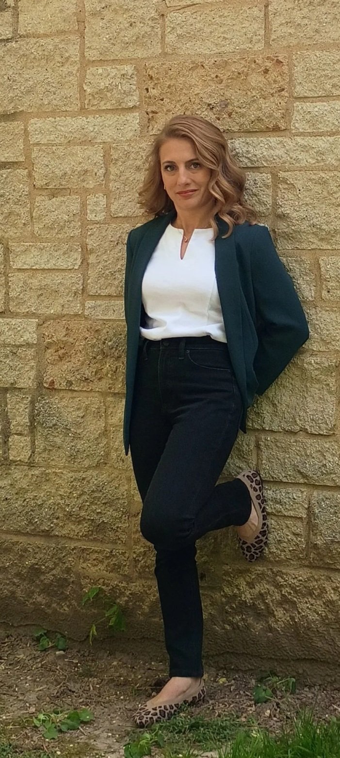 A woman with shoulder-length wavy blonde hair, wearing a teal blazer over a white top, black jeans, and leopard-print flats, standing against a beige brick wall with one foot resting against the wall.