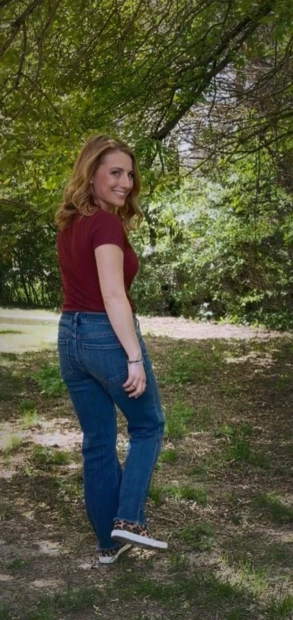 A young woman with light brown hair smiling and looking over her shoulder in a wooded outdoor area, wearing a maroon t-shirt, blue jeans, and leopard-print slip-on shoes.