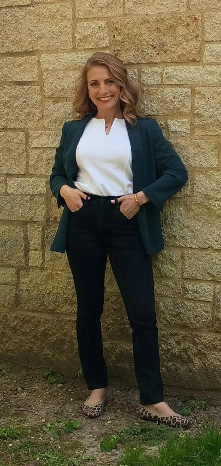 A woman with shoulder-length wavy hair smiling, wearing a white blouse, dark blazer, black pants, and leopard print flat shoes, standing against a brick wall outdoors.