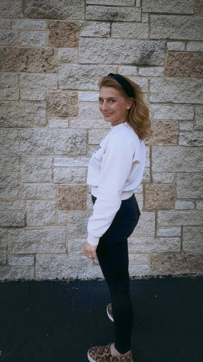 A woman with shoulder-length hair, wearing a black headband, a white sweatshirt, black jeans, and leopard-print slip-on shoes, standing against a stone wall.