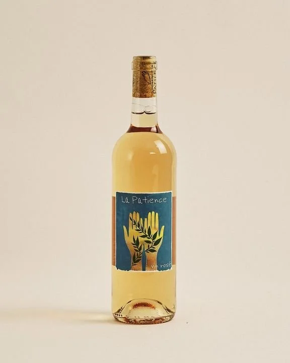 A clear glass bottle of rosé wine with a blue label that has a hand illustration and the text "La Patience."