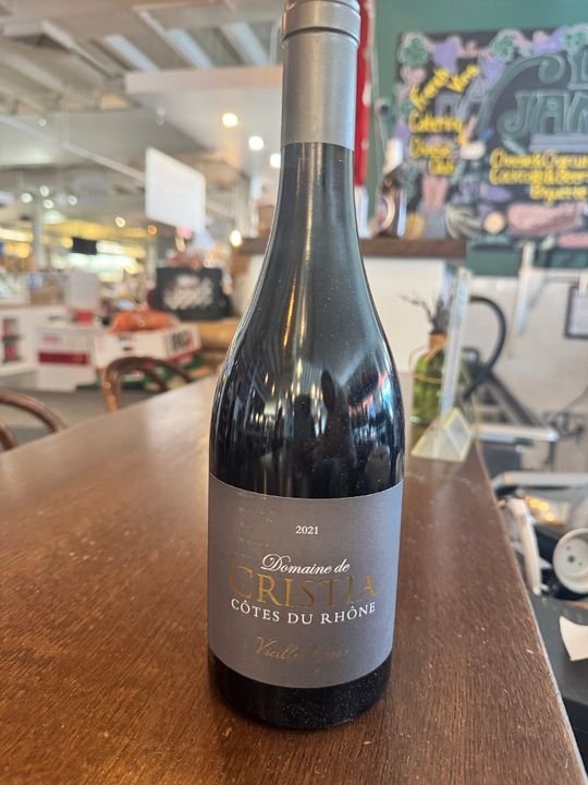 A bottle of Domaine de Cristal Côtes du Rhône wine from 2021 on a wooden table in a restaurant or cafe setting.