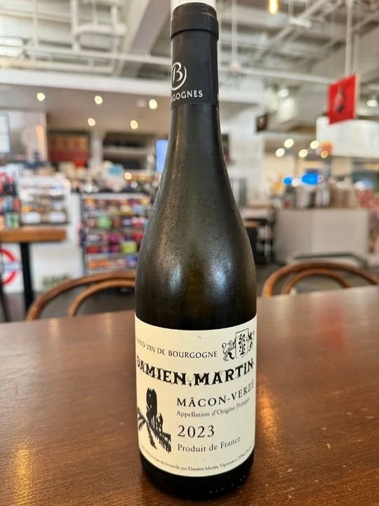 A bottle of Damién Martin Burgundy wine on a wooden table in a store or restaurant, with blurred shelves and lighting in the background.