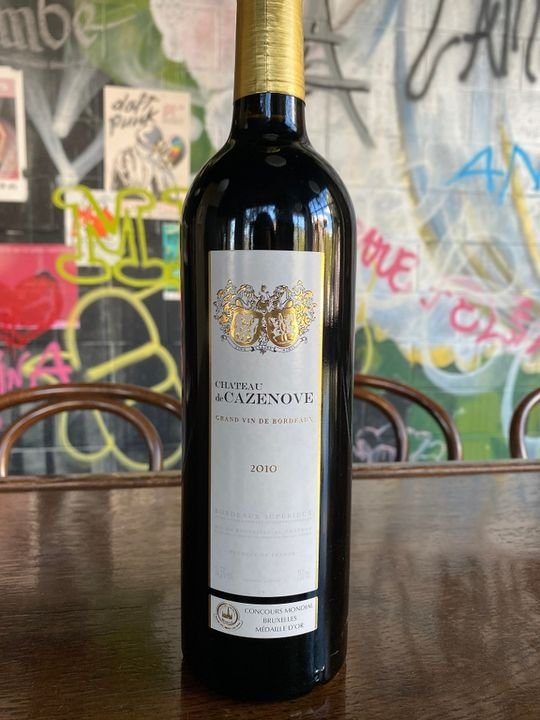 A bottle of Château de Cazenove 2010 Bordeaux red wine on a wooden table, with graffiti-covered wall in the background.