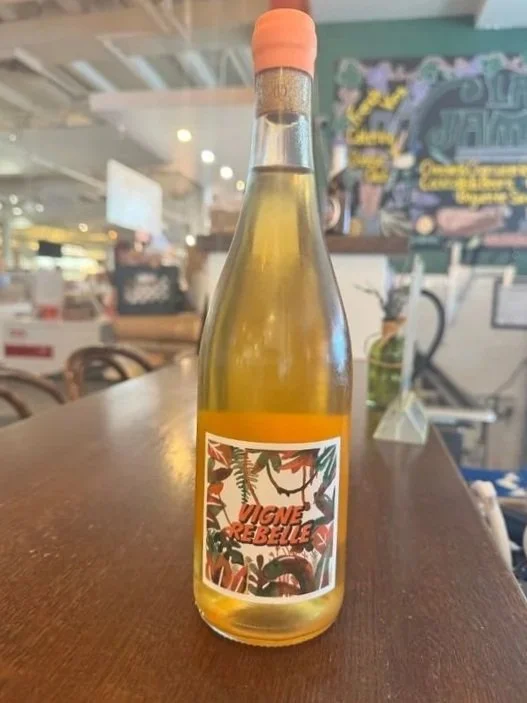 A clear glass bottle of white wine with a pink foil capsule, featuring a colorful label with tropical leaves and the text "Vigne Rebel".