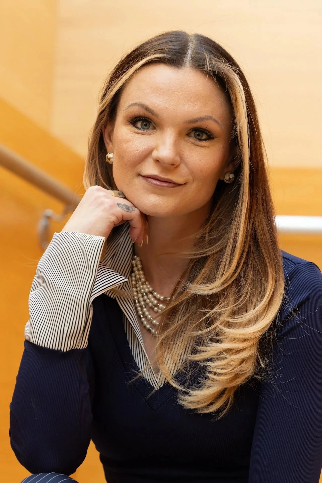 A woman with long, styled hair, wearing pearl necklaces, a navy sweater, and pearl earrings, is sitting indoors with a wooden wall and a railing in the background. She is resting her chin on her hand and looking at the camera.