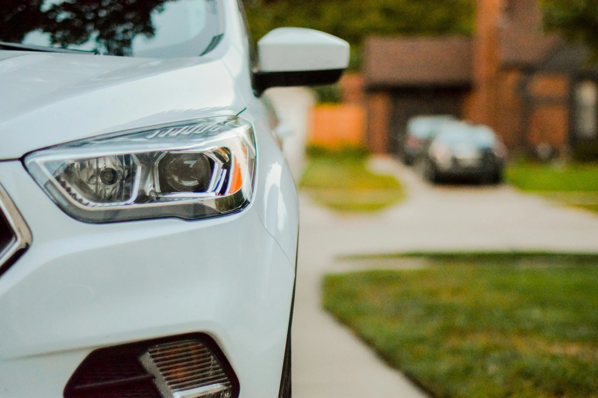 Close-up of the front part of a white pickup truck, focusing on the headlight, side mirror, and a portion of the grille, with a blurred background of cars and trees.