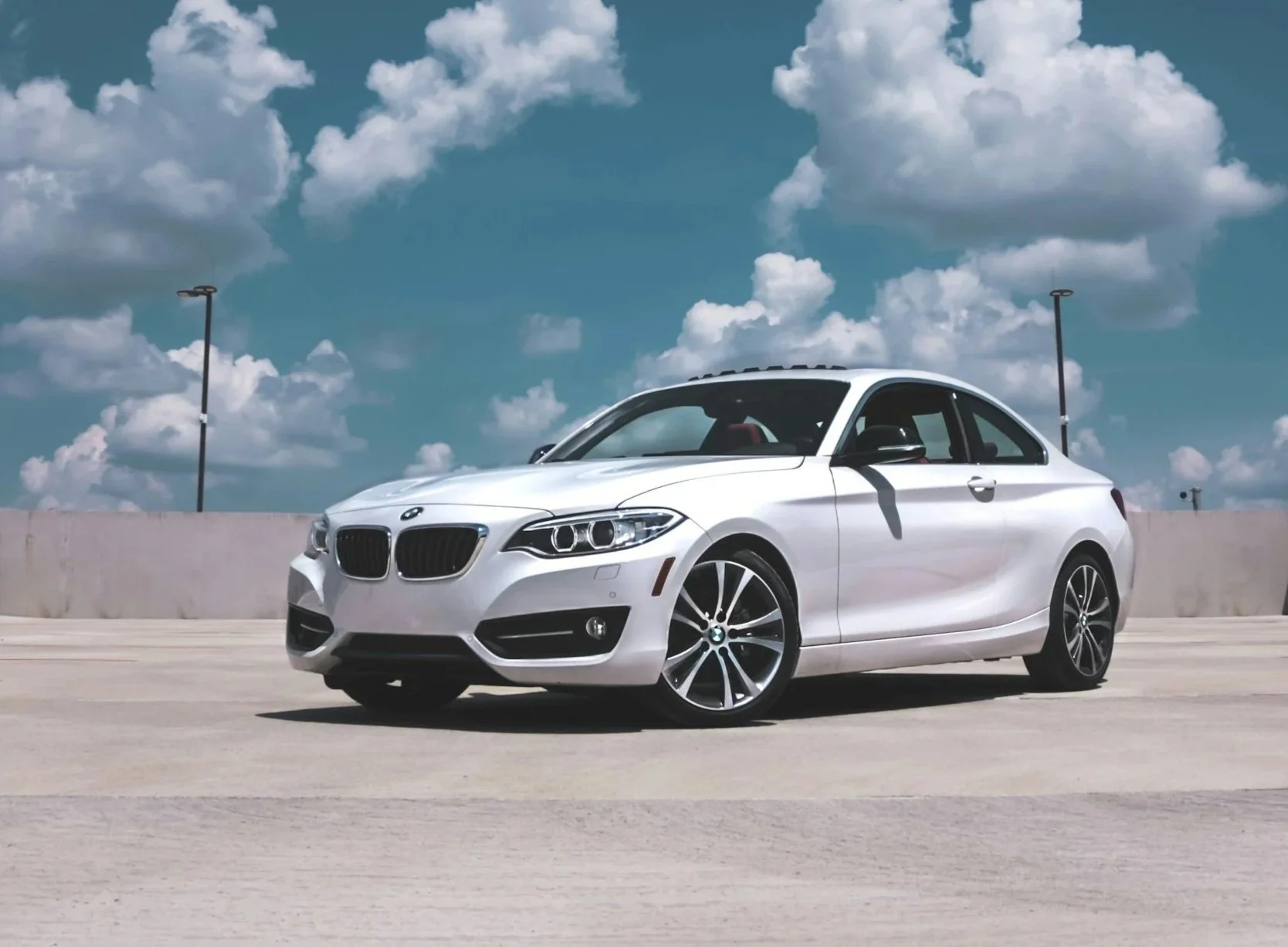 A white BMW coupe parked on a rooftop parking lot with a concrete barrier, under a partly cloudy sky with a few streetlights visible.