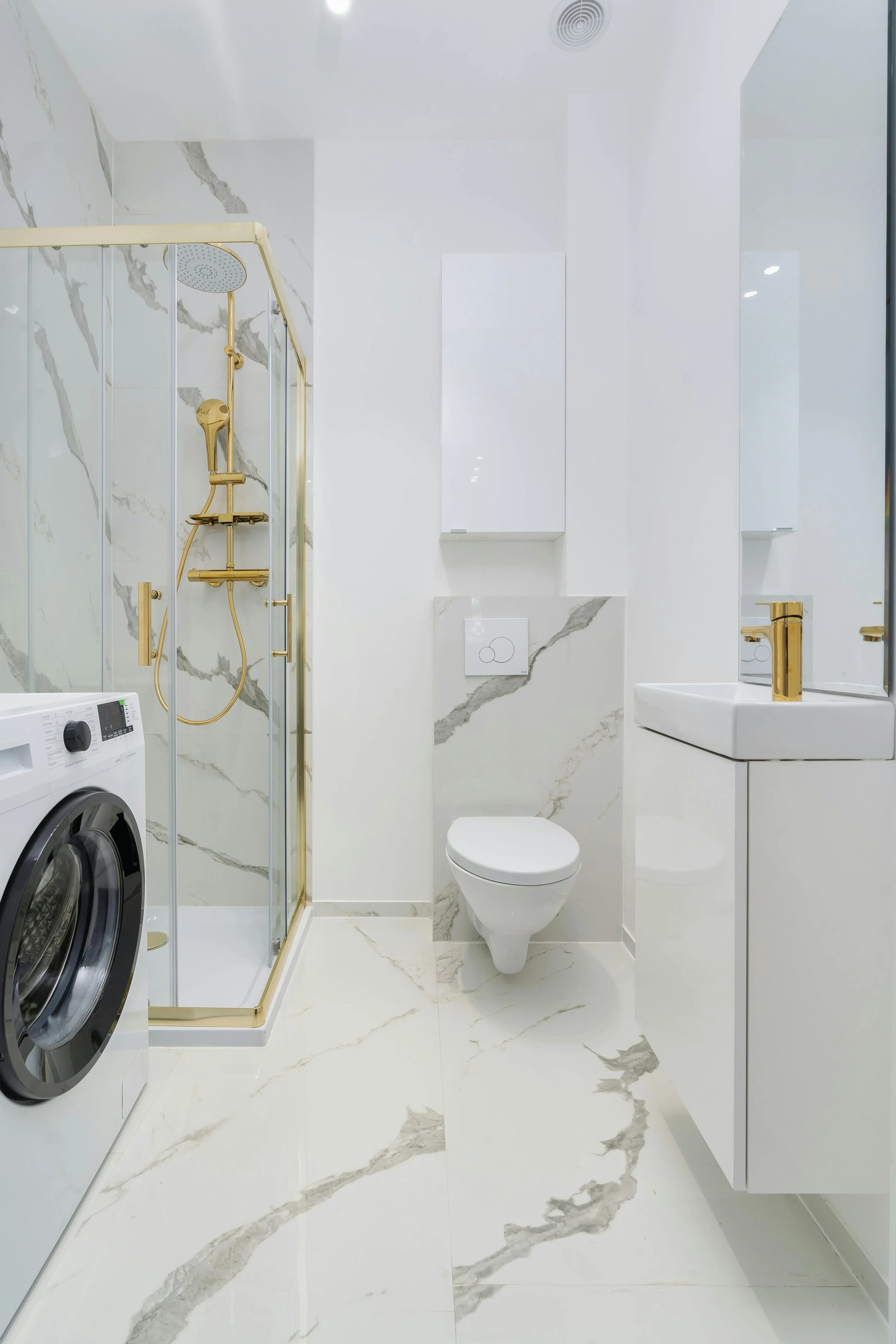 Modern bathroom with white marble tiles featuring gray veining, a glass shower with gold fixtures, a wall-mounted toilet, a white vanity with a gold faucet, and a front-loading washing machine.