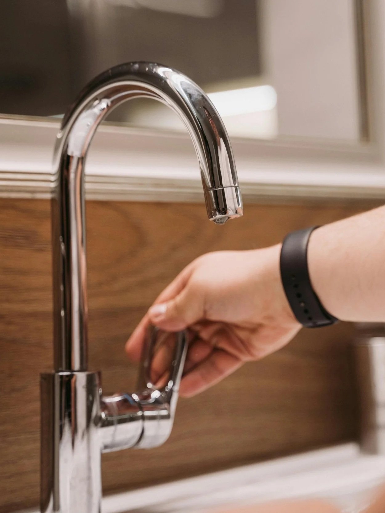 A person turning on a modern chrome kitchen faucet with their right hand, wearing a black wristband, over a wood-textured kitchen counter.