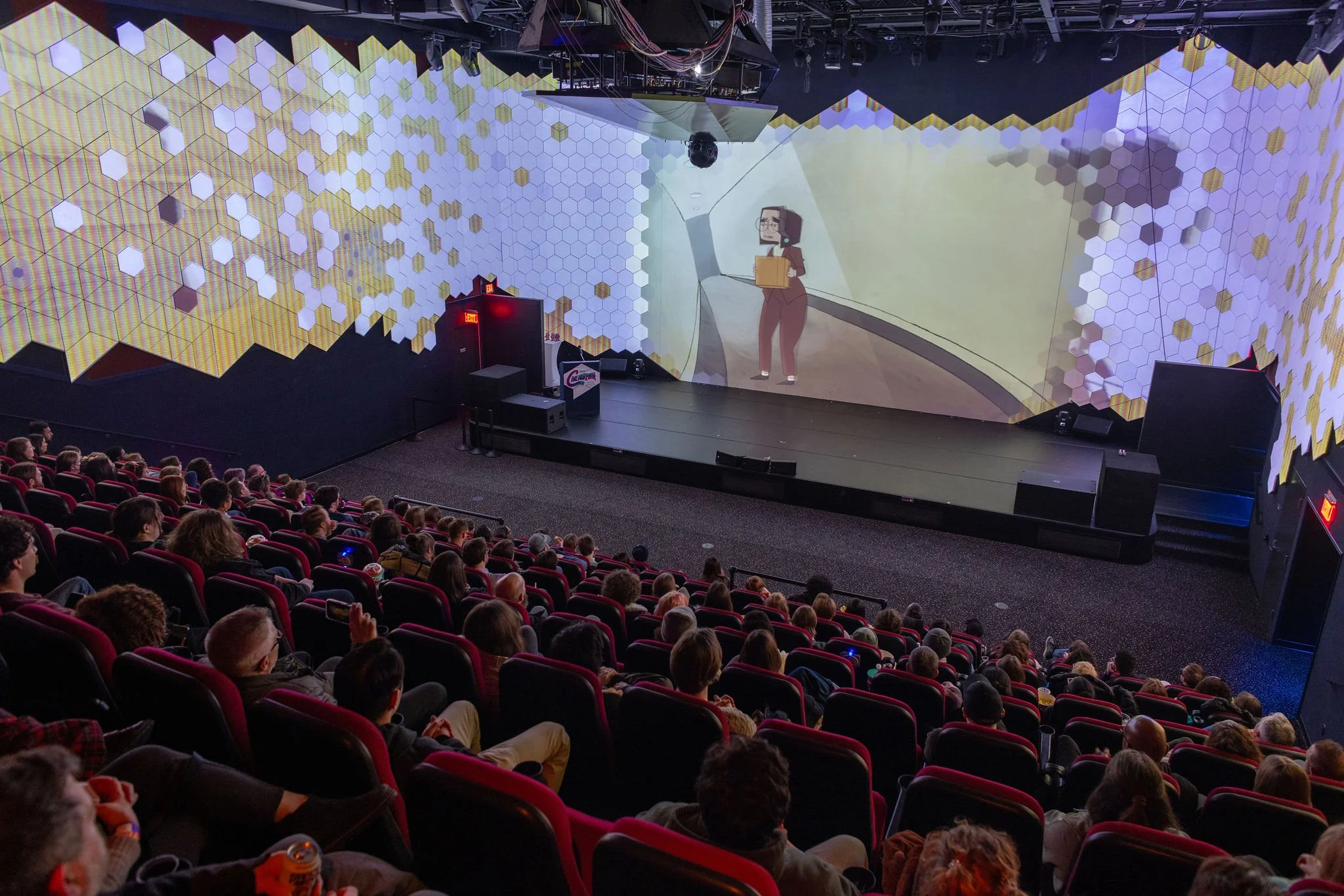 A large theater with a stage and audience, showing a projection of an animated woman holding a box on the screen, with decorative honeycomb-like wall designs inside Atlas9.