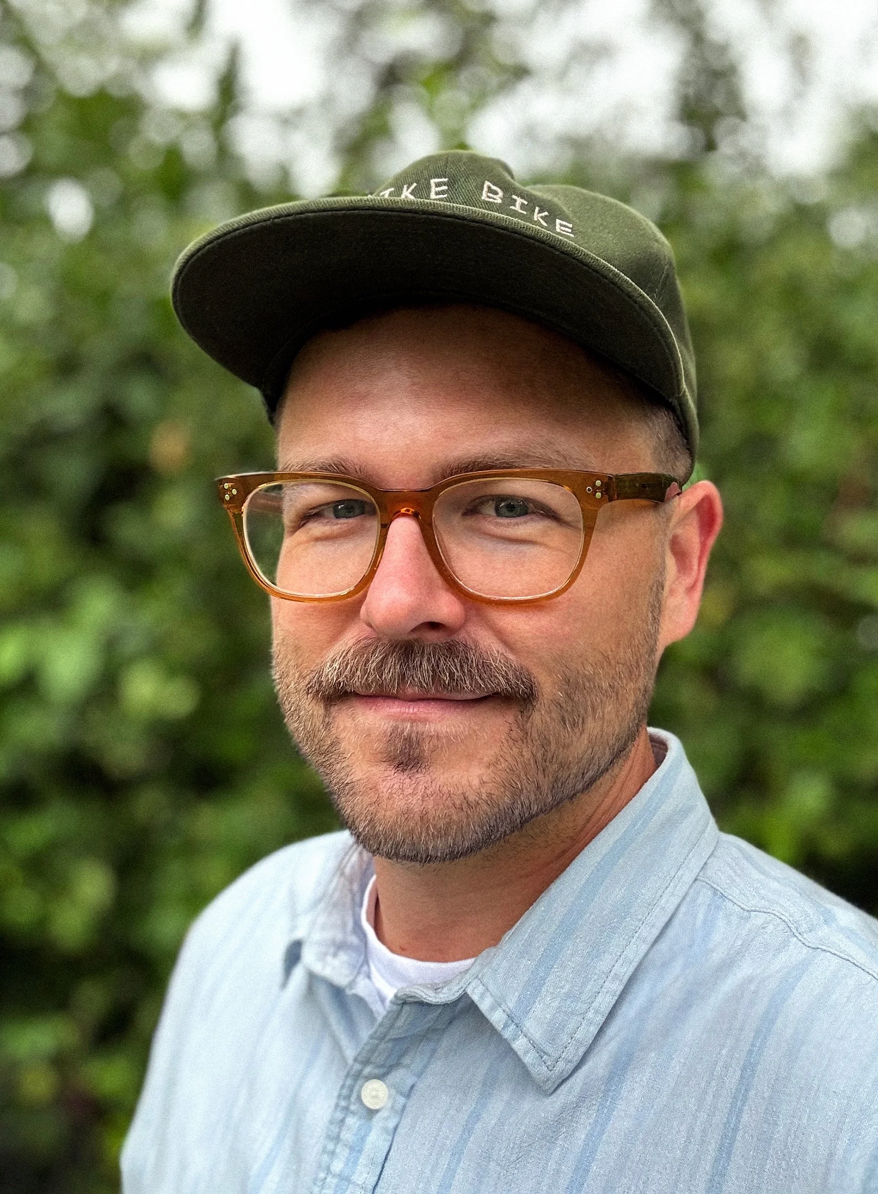 Close-up of a man with light skin, wearing glasses and a blue striped button-up shirt, outdoors with blurred greenery in the background.