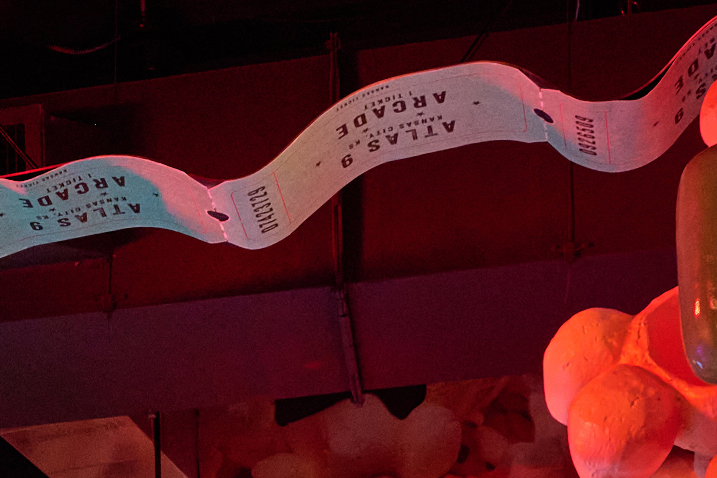A paper movie ticket with the words 'ATLAS'S 9 ARCADER' hanging from the ceiling in a dimly lit room with red lighting inside Atlas9.