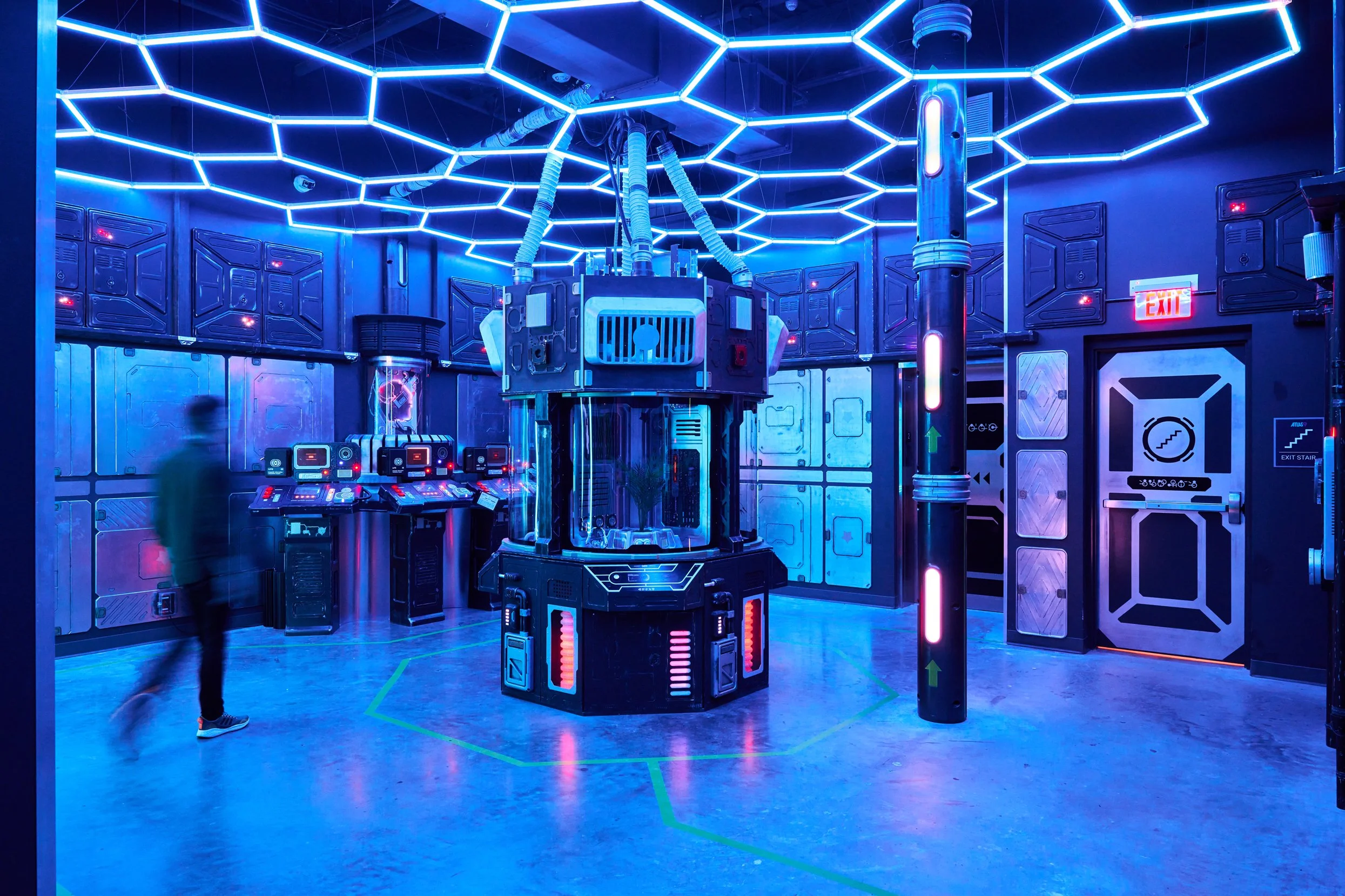 A futuristic, neon-lit room with blue lighting, high-tech equipment, and a person walking by.