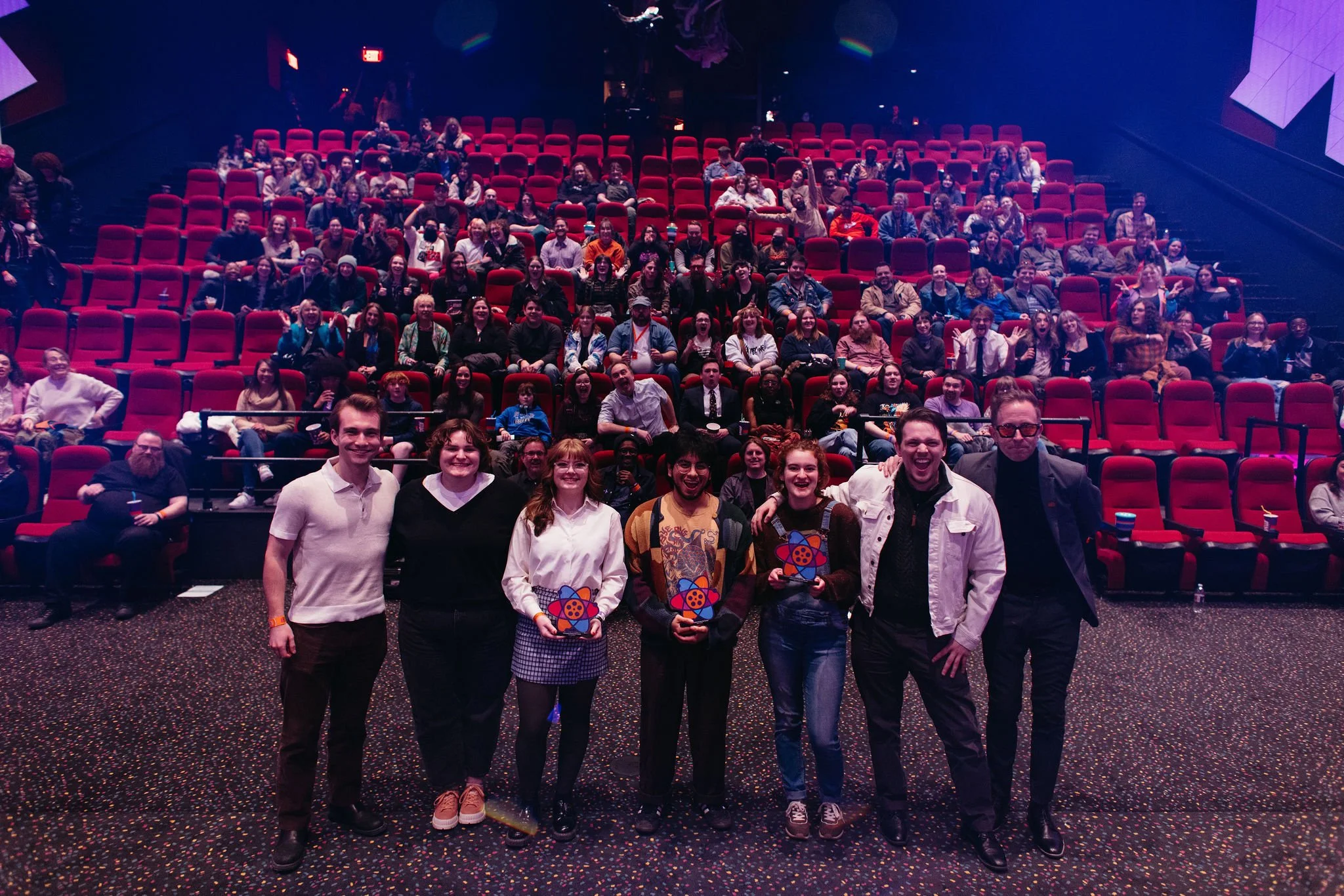Group of seven people standing in front of an audience inside Atlas9 inside a theater or auditorium, with many rows of red seats filled with spectators in the background.