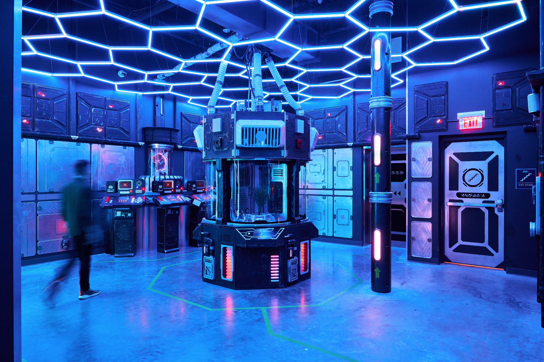 A futuristic room with blue lighting, featuring a central sci-fi device with tubes, control panels on the sides, and geometric lighting on the ceiling. A blurred person stands nearby, and there are various panels, doors, and an exit sign.