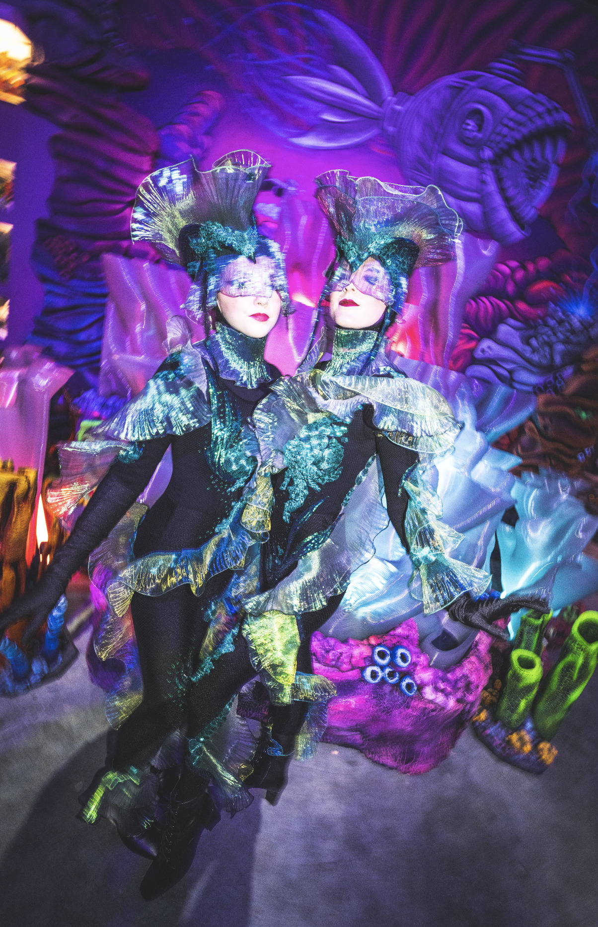 Two women performers from Quixotic dressed in elaborate, luminous, fish-inspired costumes with headpieces, posing in front of a vibrant, abstract aquatic backdrop inside of Atlas9.