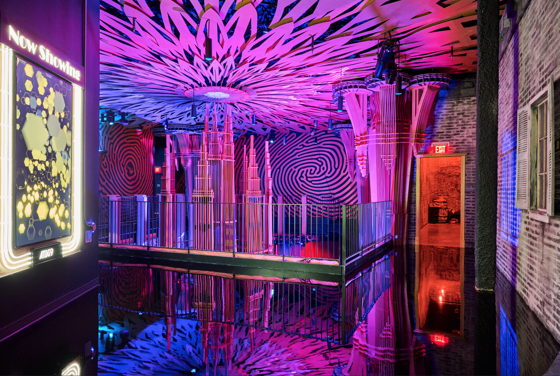 An interior space with vibrant, colorful neon lighting, featuring a modern art installation with pink, purple, and blue hues. The ceiling has an abstract, floral-like design, and the walls display swirling patterns. Reflection on a glossy floor enhan
