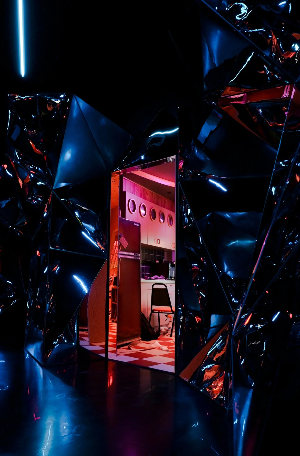 View of a colorful, reflective, and darkly lit corridor inside of Atlas9 with a door frame leading to a kitchen with pink and purple neon lighting. The kitchen has checkered flooring, a refrigerator, and chairs.