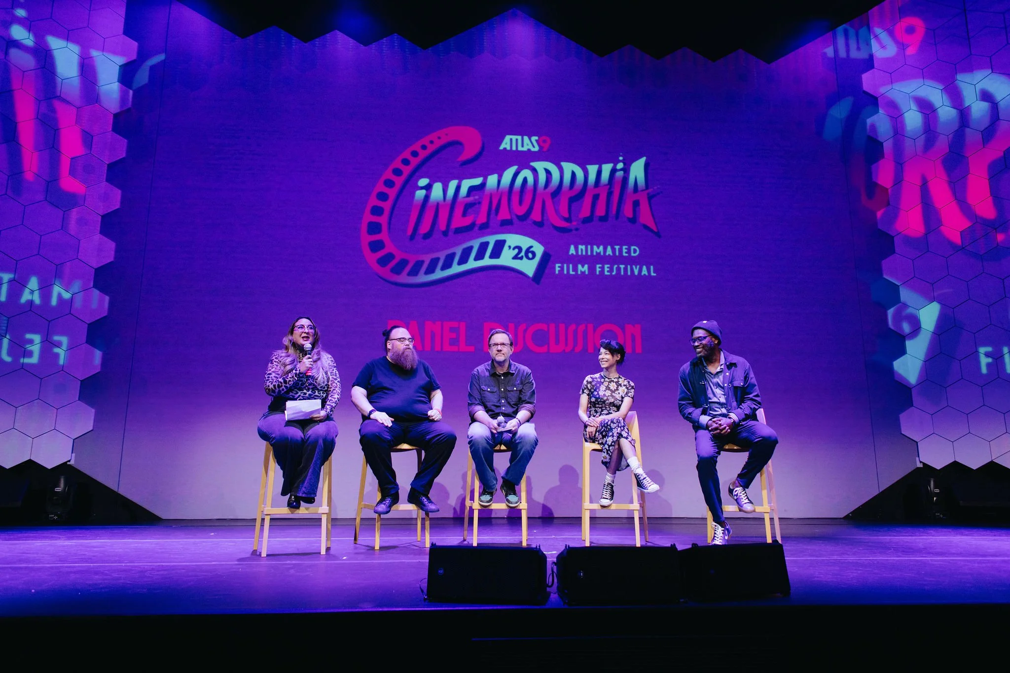 Panel discussion on stage at the Atlas 9 Cinemorphia Animated Film Festival, with five people seated on high chairs and a large screen behind them displaying event graphics.