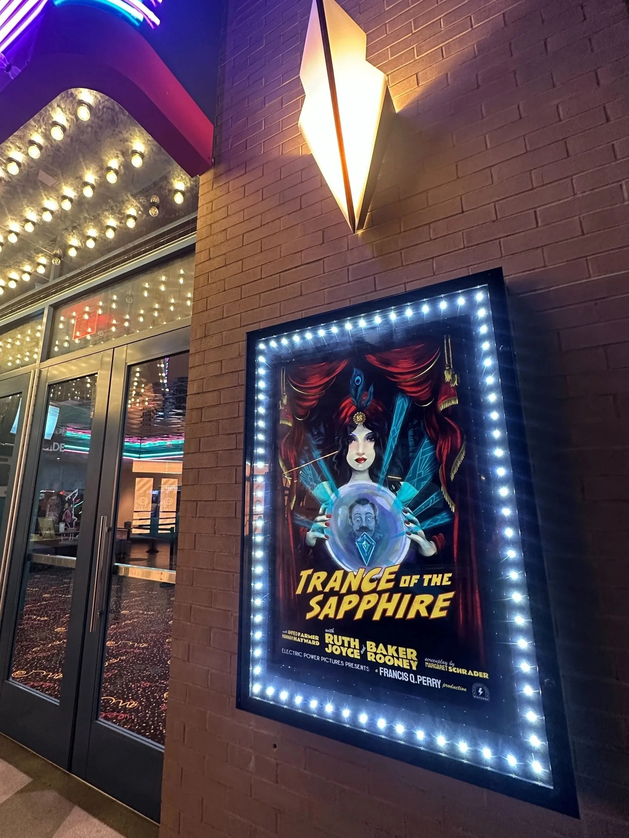 Poster for the show "Trance of the Sapphire" featuring an illustration of a woman with dark hair, wearing a feathered headpiece, holding a glowing crystal ball with a man's face inside, framed by a border of lights, outside the theater entrance.