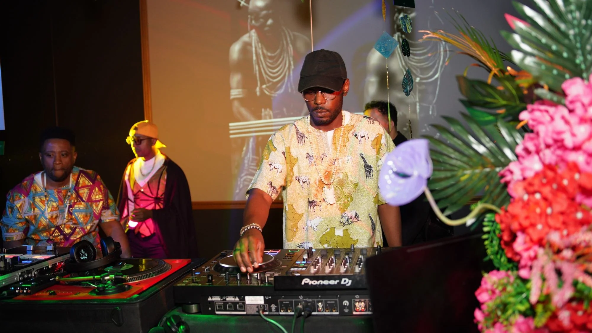 A DJ wearing a black cap, glasses, and a patterned yellow shirt with animal prints, is operating DJ equipment at a party or event. Behind him, two people are visible; one in a colorful patterned shirt and another with a hoodie and glasses. Bright pin