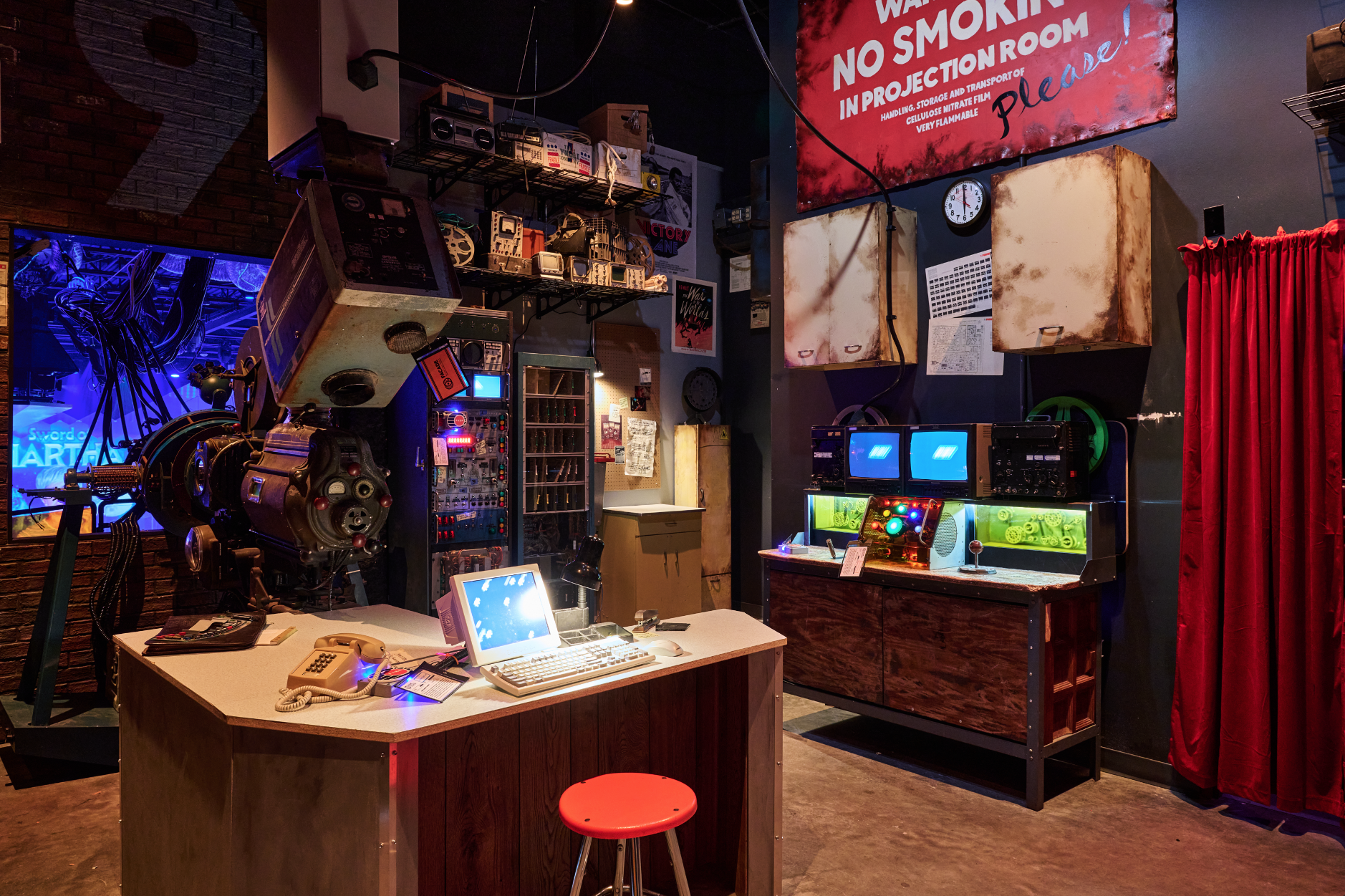 A vintage projection room with various outdated electronic equipment, including a large film projector, monitors, and control panels. A red curtain is on the right, and a large warning sign about no smoking is hanging on the wall. The room is clutter