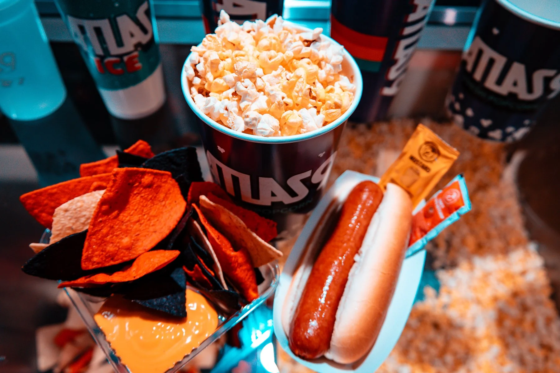 A tray of nachos with cheese, a hot dog with ketchup, a cup of popcorn, and several cups of soda all branded with Atlas9 logos. The food is from Atlas9 concessions