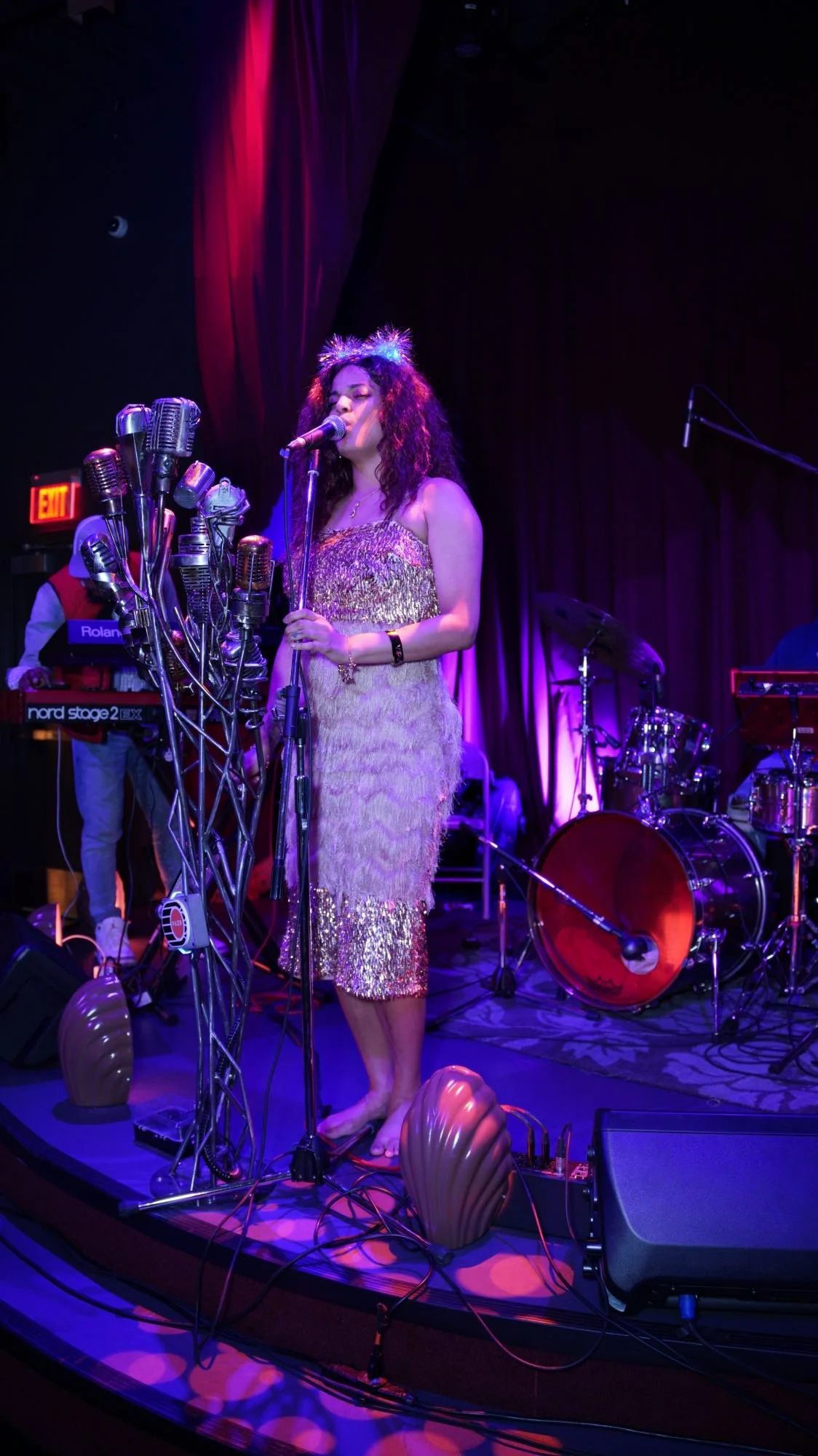 A female singer on stage in the jazz club at Atlas9 with vintage microphones, standing barefoot in a glittering dress, with a band setup including drums and keyboard behind her, illuminated by purple and pink stage lighting.