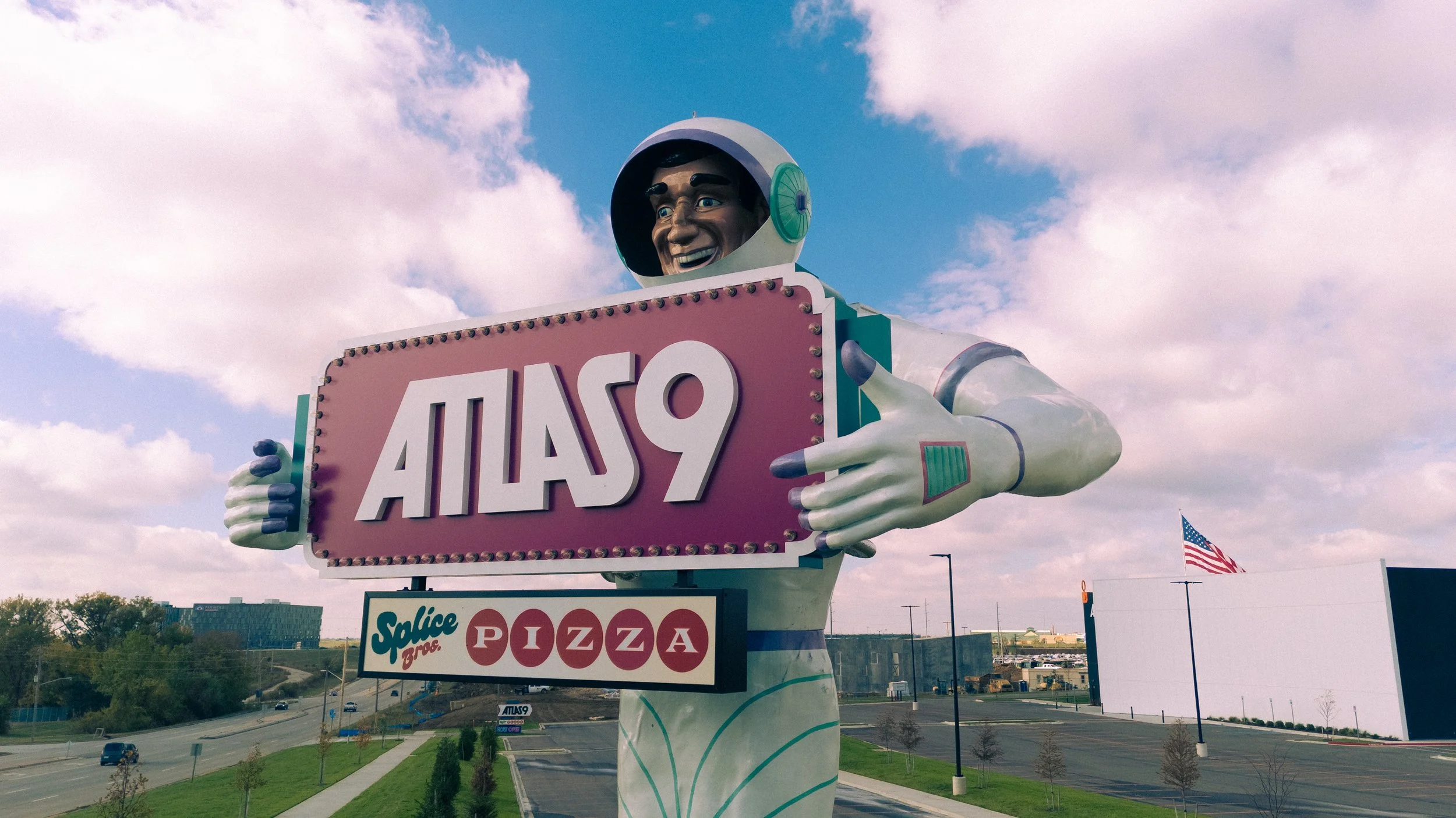 A large retro-style sign in the shape of an astronaut holding a pink sign that says 'ATLAS9', with a smaller sign below advertising 'Slice Bros Pizza'. The scene is set outdoors on a partly cloudy day with an American flag in the background.