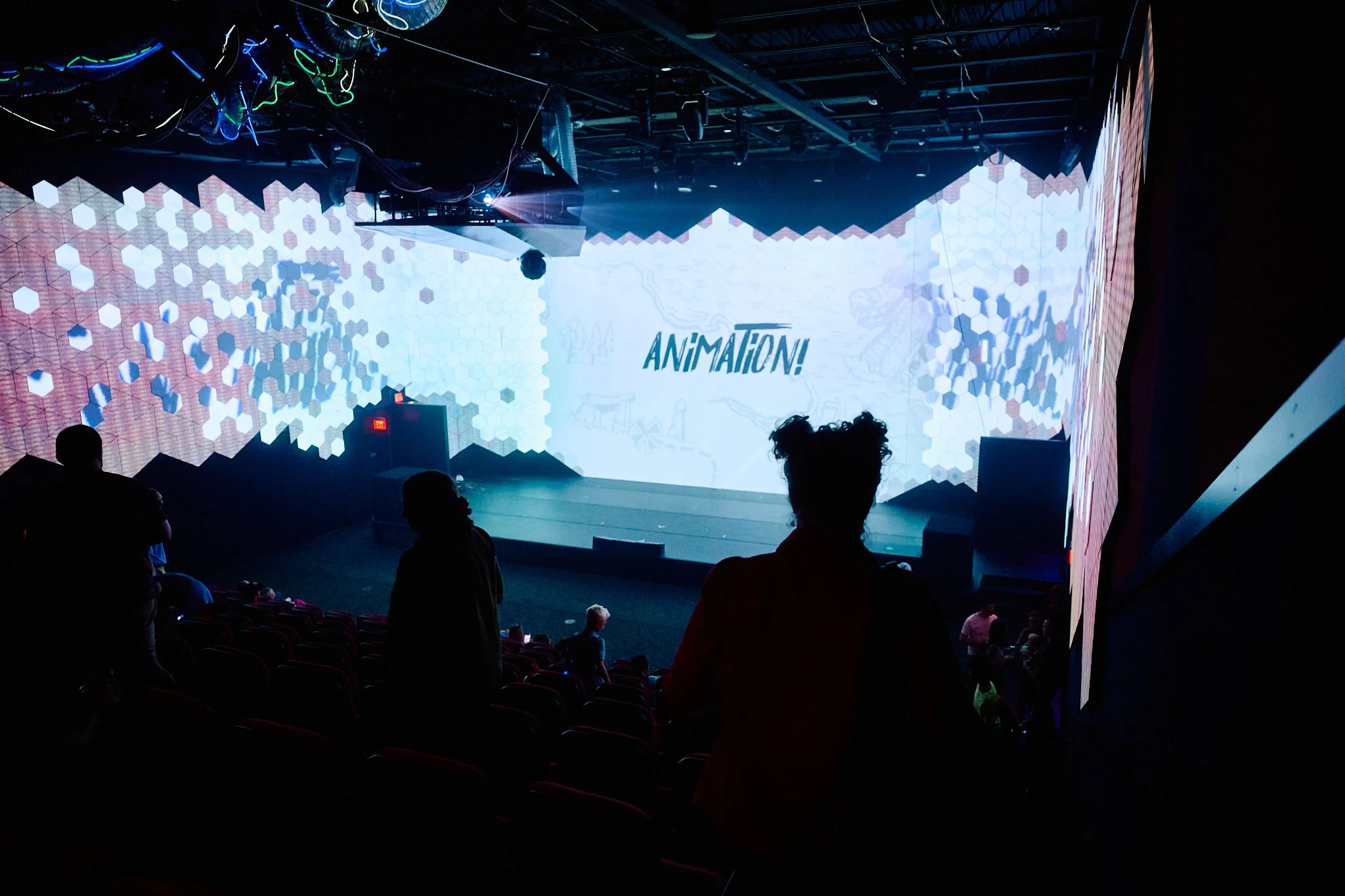 The image shows a dark theater or auditorium with silhouettes of people standing and sitting in front of a stage. The stage has a large LED screen displaying the word 'ANIMATION!' and a background with hexagonal patterns, with some abstract sketches 