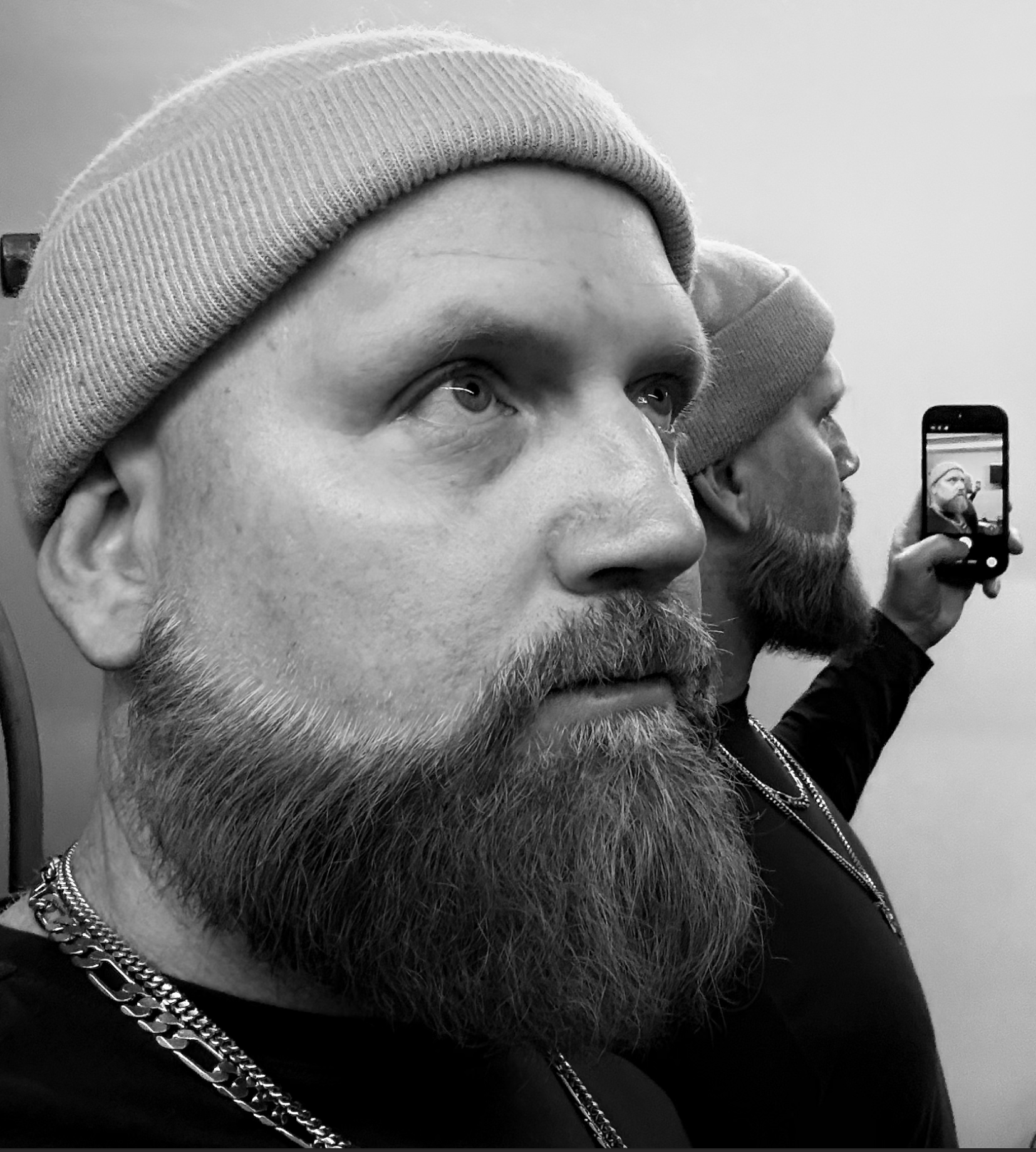 Close-up black-and-white photo of a man with a beard and light beanie hat, with another person in the background holding a smartphone capturing the same shot.