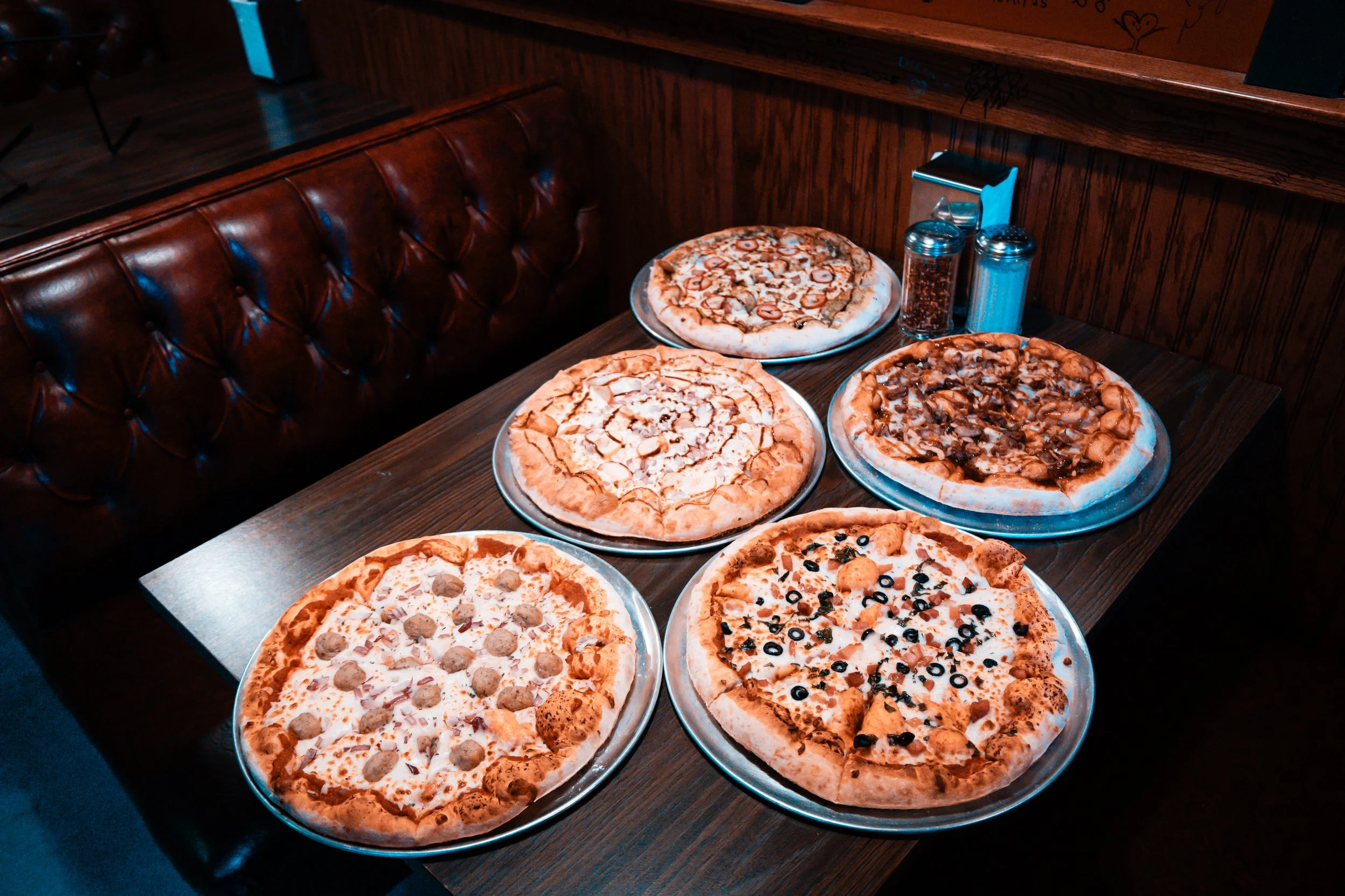 Six pizzas from Splice Bros. Pizza in Atlas9 on metal trays on a wooden table in a restaurant with a leather booth seat and condiment shakers.