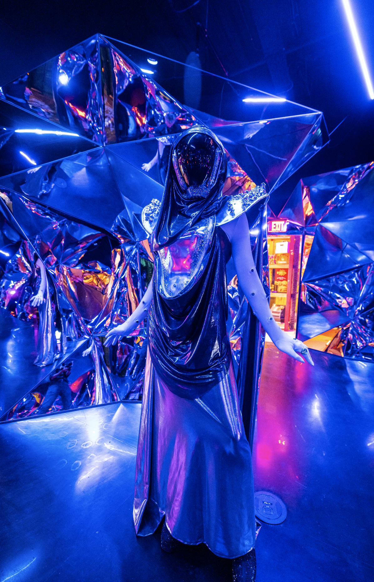 A Quixotic performer dressed in a metallic, dark, futuristic costume with a large hood, set against a background of reflective, prism-shaped surfaces illuminated with blue, purple, and pink neon lights at Atlas9.