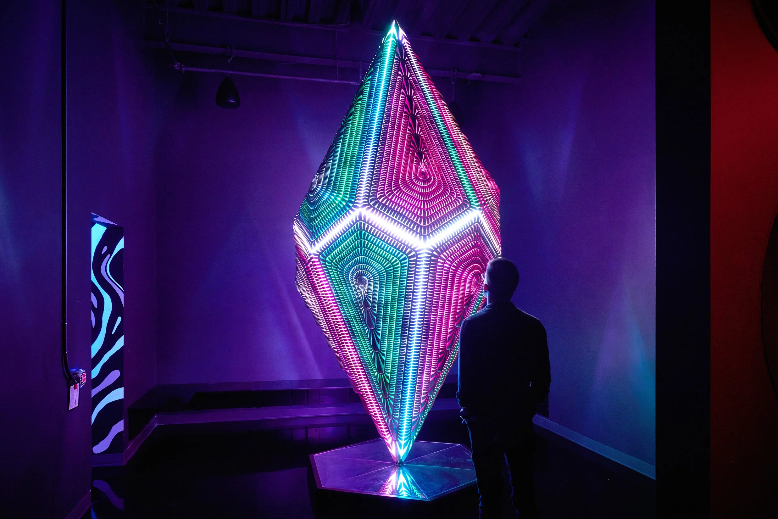 A person standing in front of a large, illuminated, colorful geometric sculpture in a dark room. The sculpture is a diamond-shaped object made of numerous small, glowing LED lights arranged in intricate patterns.
