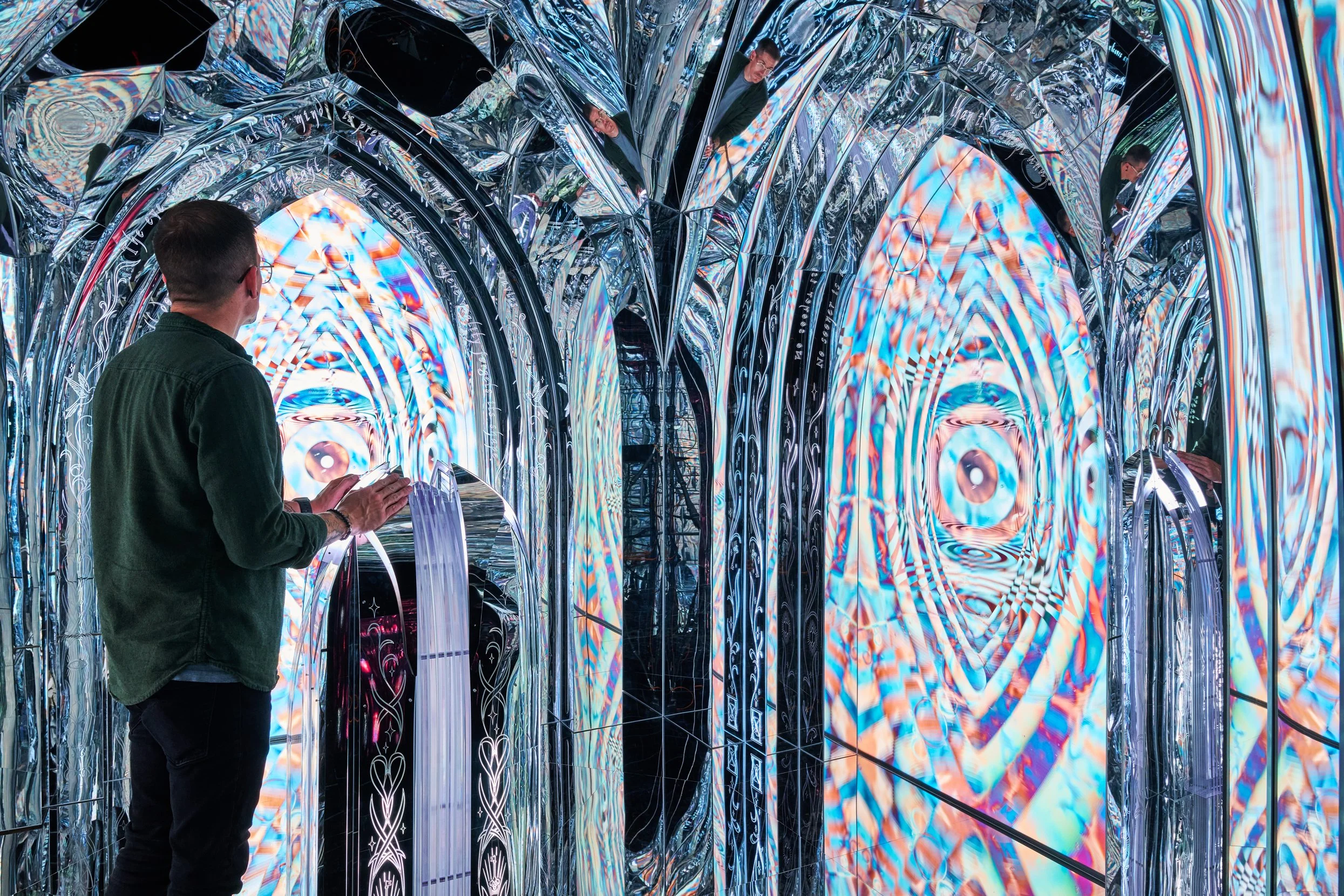 A man in a green jacket is standing inside an immersive art installation with reflective and metallic surfaces, holding a smartphone. The installation features arched, tunnel-like structures with colorful, kaleidoscopic digital projections and reflec