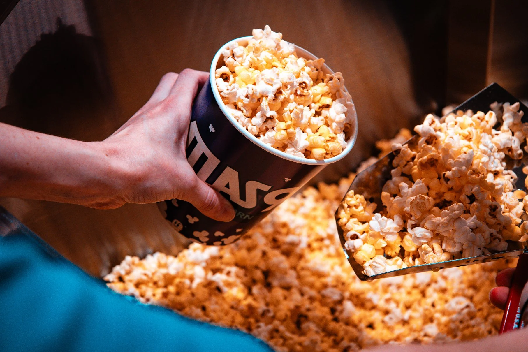 Person scooping popcorn from the Atlas9 concession stand while holding a paper bucket of popcorn with an Atlas9 logo. 