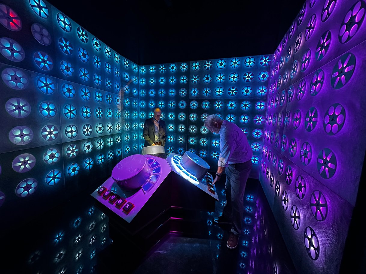 Two men interacting with a futuristic, illuminated control panel inside a room with walls covered in glowing, circular patterns of blue and purple lights.