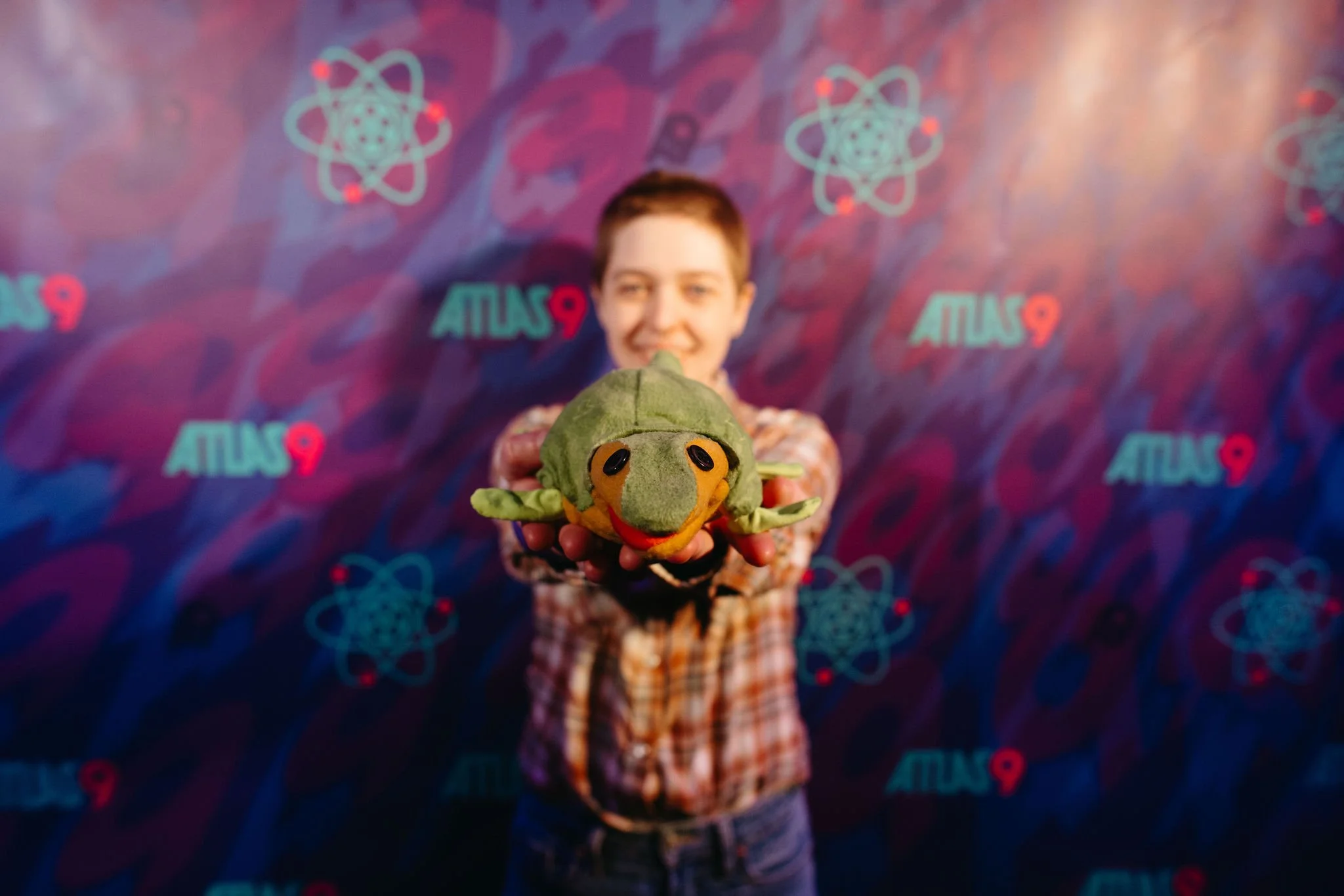 A person holding a plush turtle toy in front of a colorful background with the words 'Atlas 9' and science-themed icons.