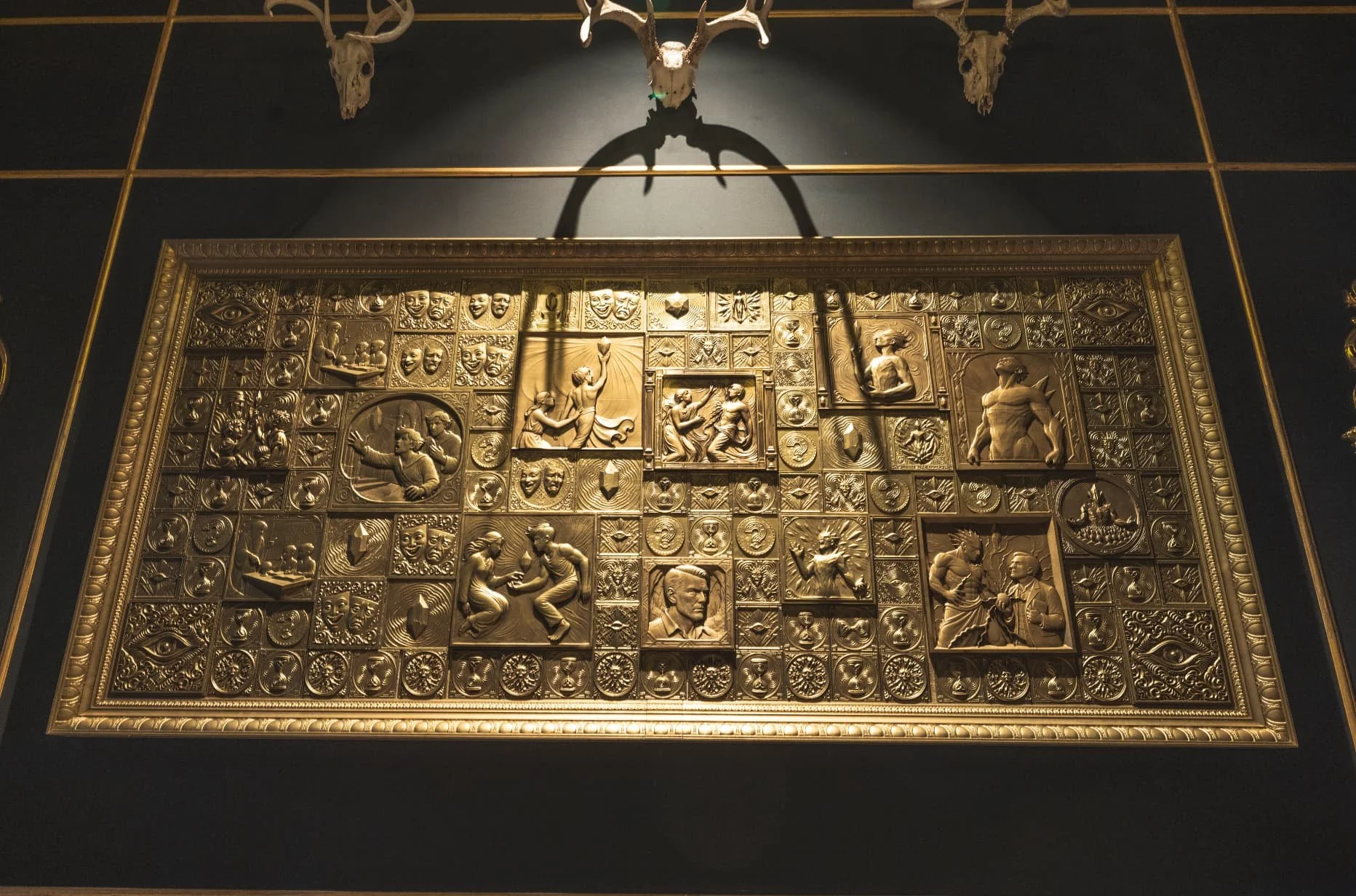 A large, ornate gold plaque with detailed embossed images and portraits, mounted on a dark wall with a skull and antlers mounted above it at Atlas9. The plaque features various scenes and faces in a grid pattern.