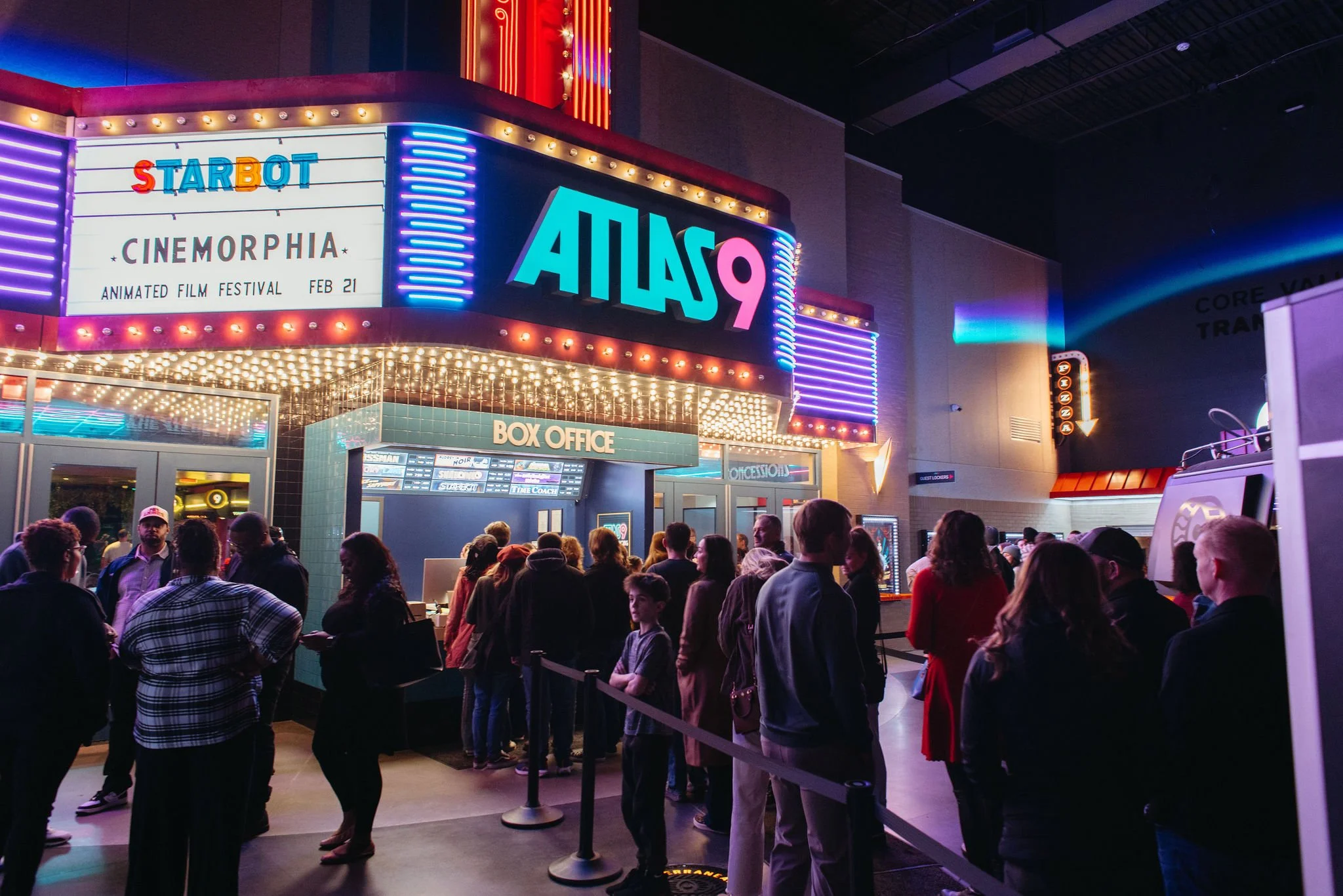 Line of people waiting outside a movie theater at night with neon lights and marquee signs, including 'Cinemorphia' and the theater name 'Atlas9'.